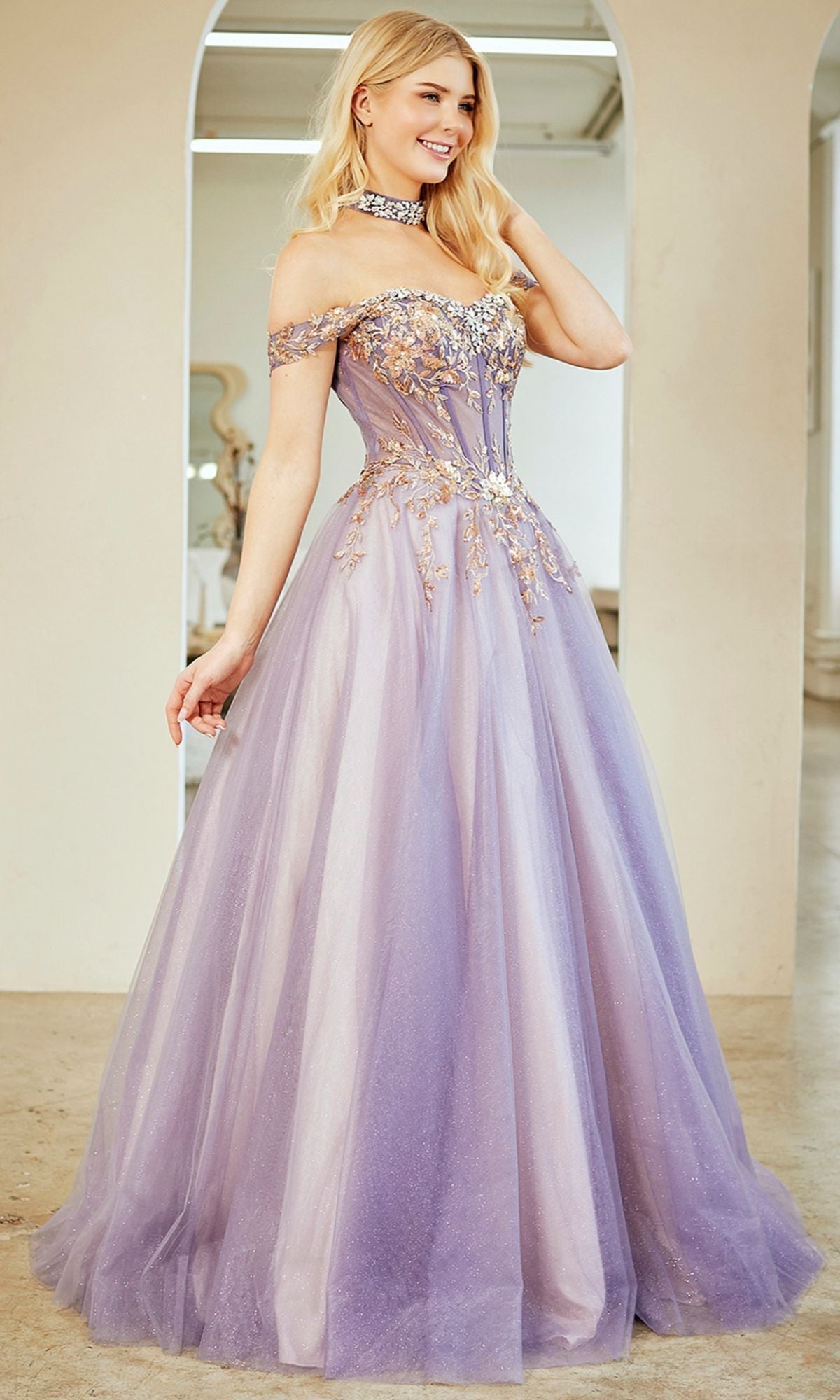 A woman stands indoors wearing the Amelia Couture 8063, a long prom dress in lilac with gold detailing, sequin appliquรฉ, an off-the-shoulder neckline, and a matching choker.